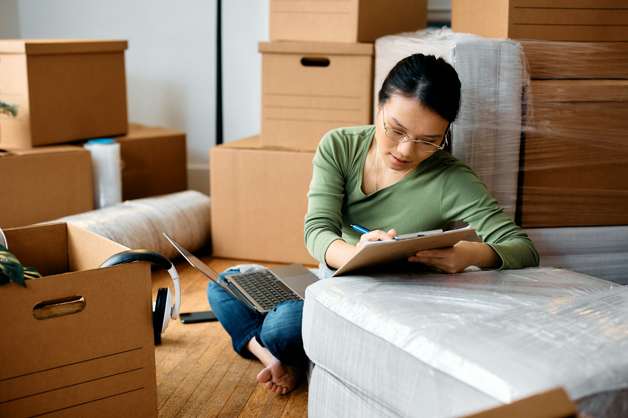 Asian woman going through checklist while using laptop and moving into a new home.