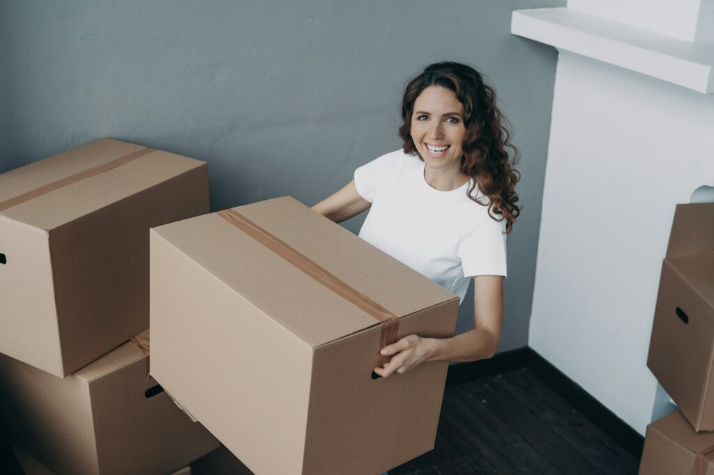 Joyful female homeowner standing in new house holding cardboard box. Moving, apartment renovation